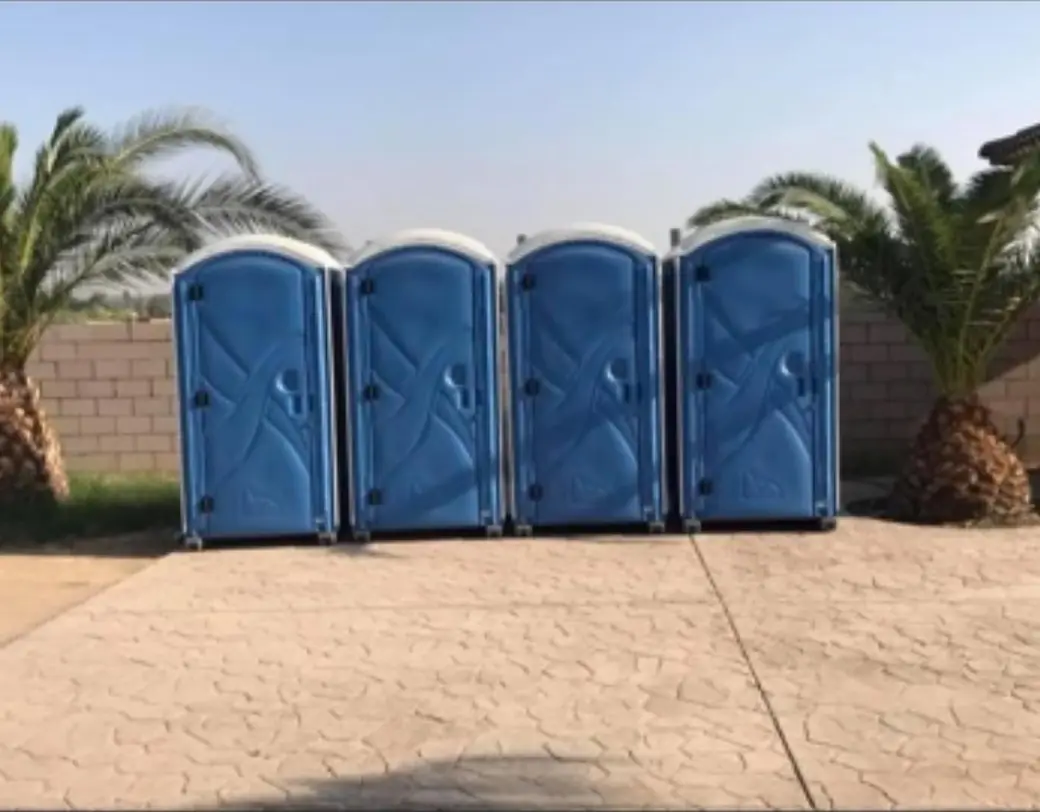 Four blue portable toilets in a line on a stamped-concrete patio with palm trees