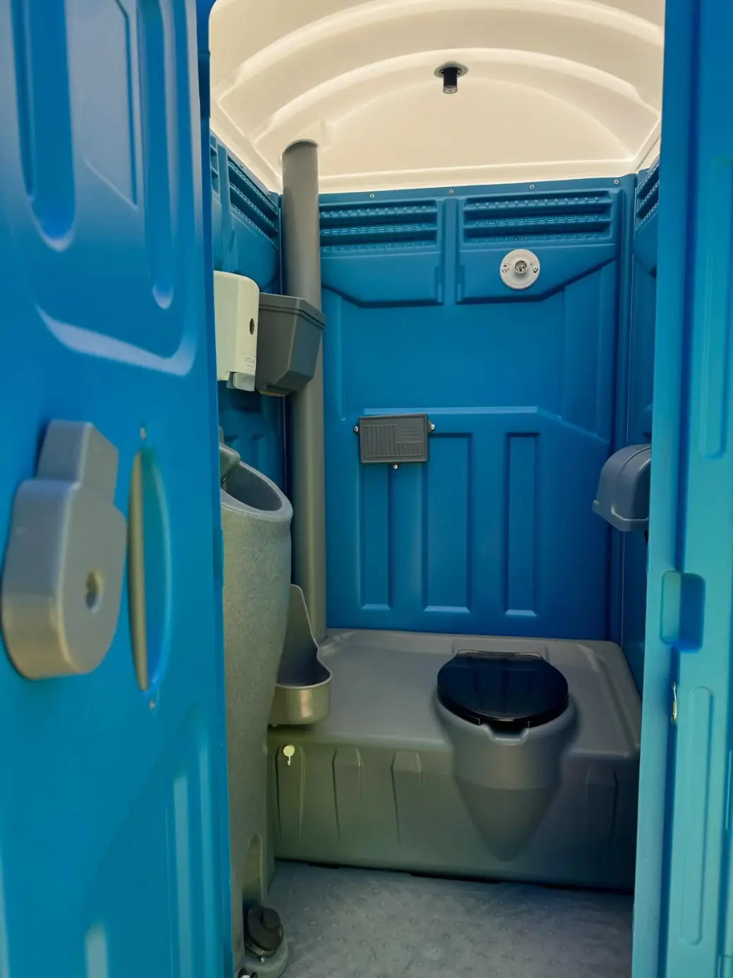Inside a blue portable toilet showing the seat, urinal, vent pipe and paper holder