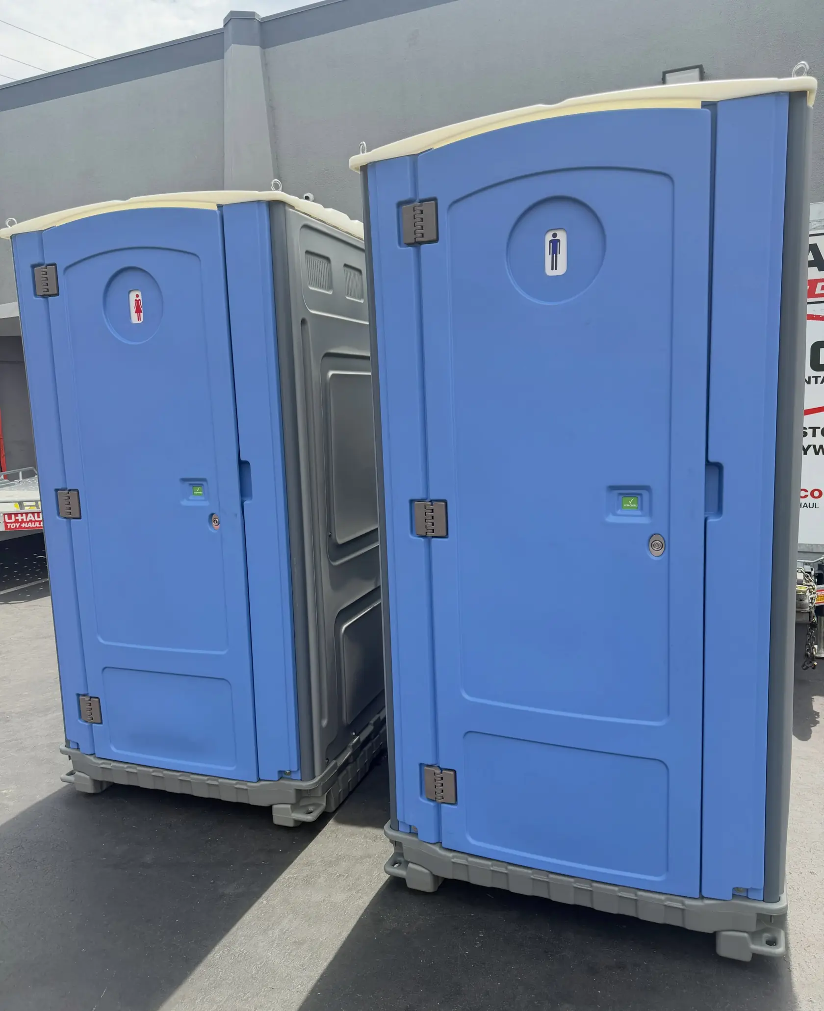 Two blue portable toilets on asphalt beside a metal commercial building