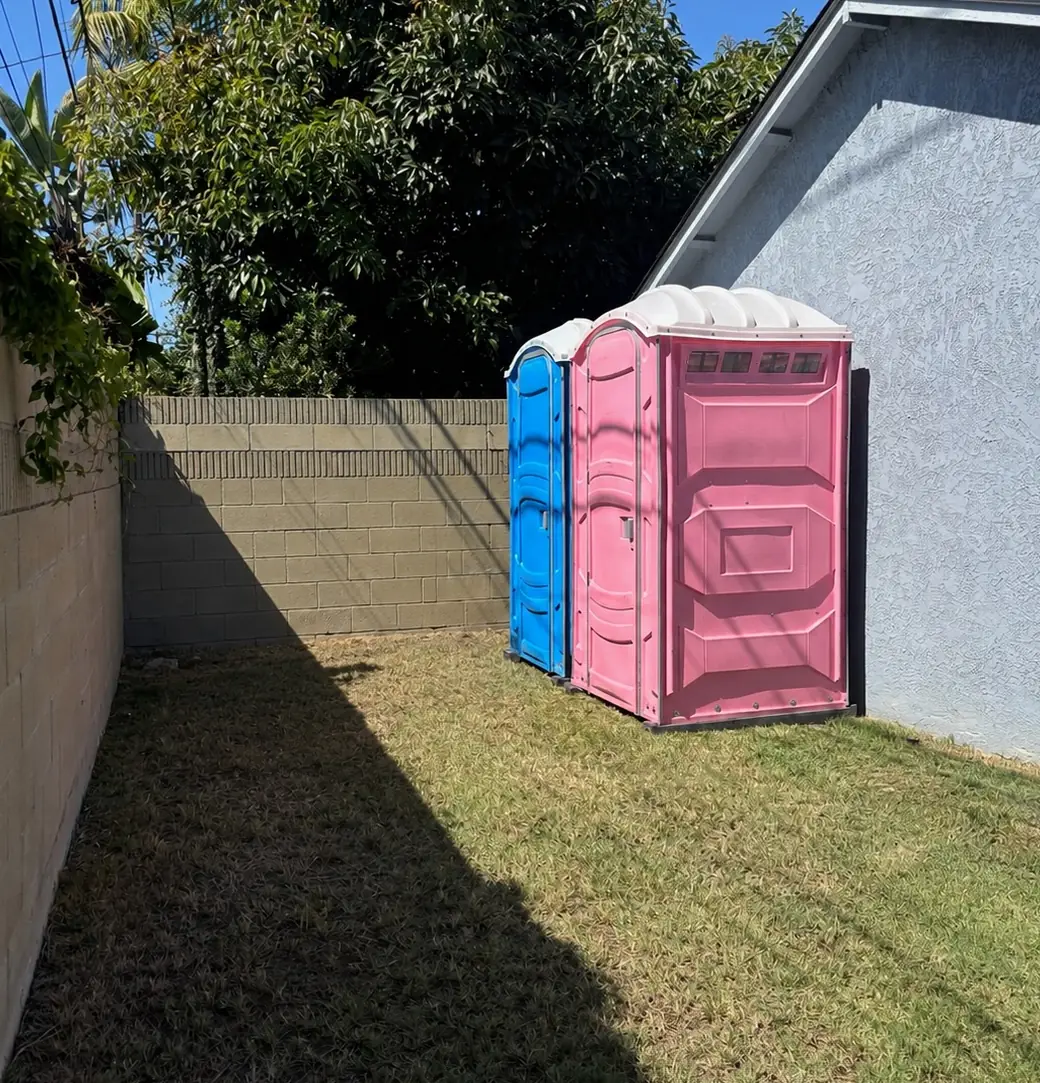A blue and a pink portable toilet set on grass inside a fenced backyard