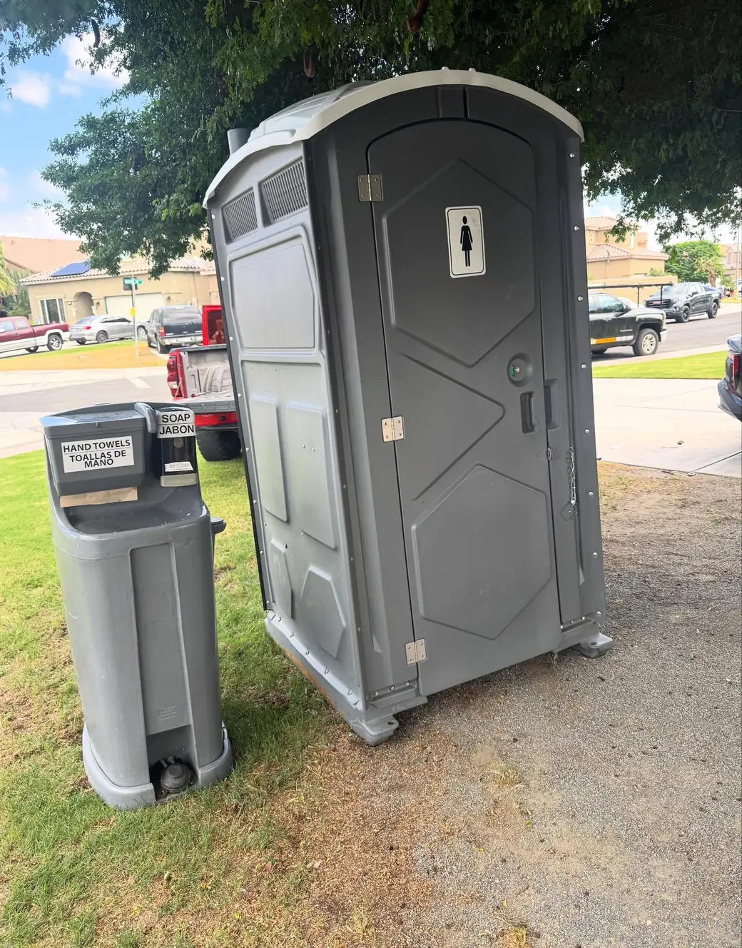 Single gray portable toilet marked for women beside a foot-pump hand-wash sink on grass