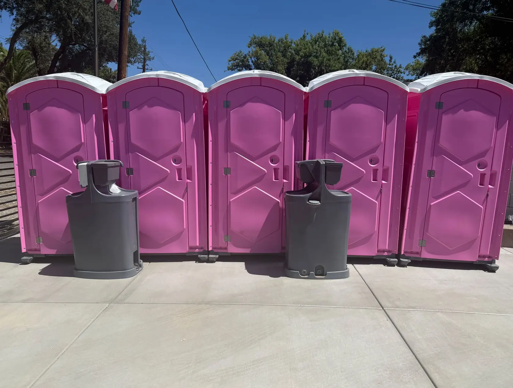 Five pink portable toilets lined up with two gray hand-wash sinks in front
