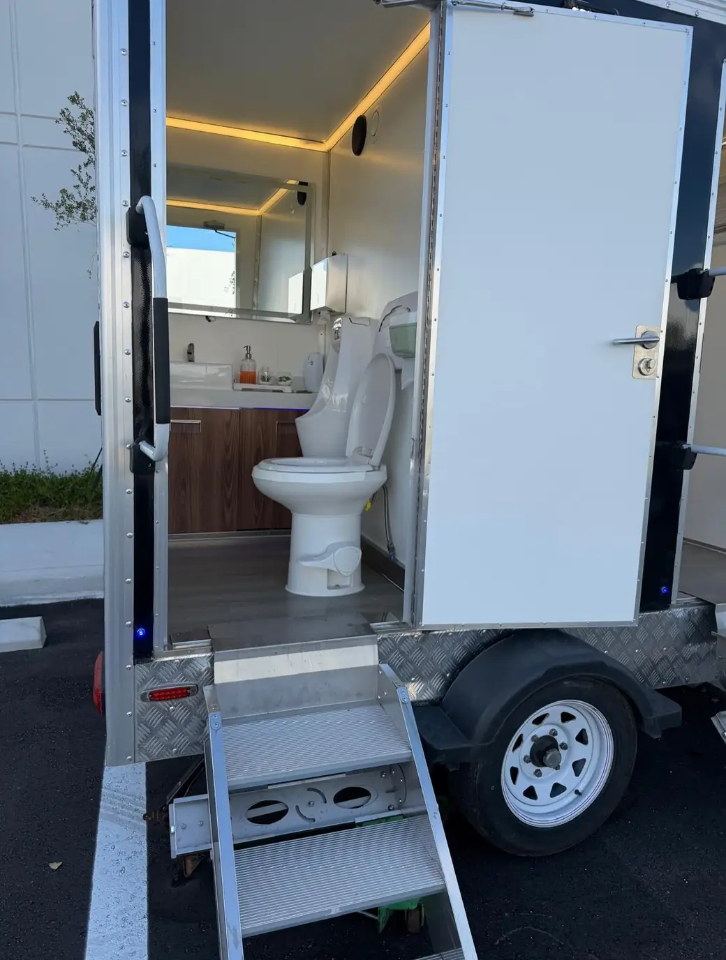 Open restroom trailer stall with a flushing toilet, wood vanity and lit mirror