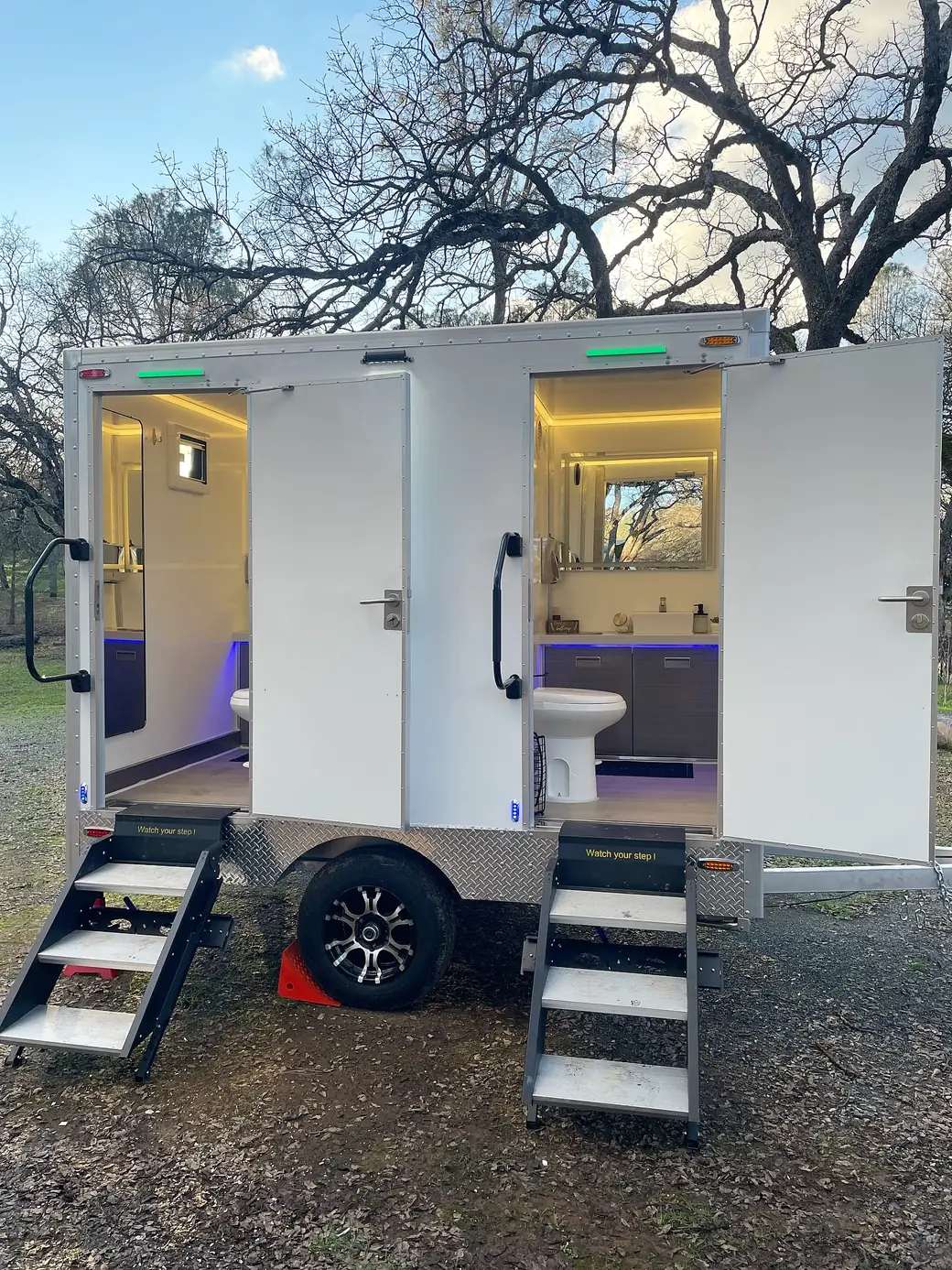 Two-stall restroom trailer with lights on and doors open at an evening event